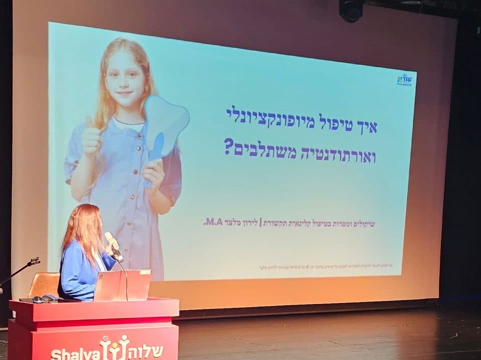 לירון מלצר מרצה על במה ליד דוכן. על המסך: איך טיפול מיופונקציונלי ואורתודונטיה משתלבים?